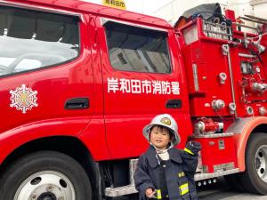 消防車の前で、防火服とヘルメットを被った幼稚園児の女の子