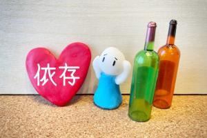 依存の文字と頭を抱えて困った顔の人形とアルコールの瓶の写真