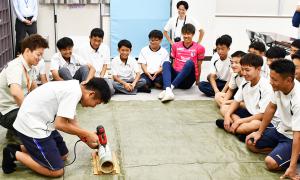 ドリルで穴を開ける男子生徒とそれを笑顔で見守る田中選手と生徒たち