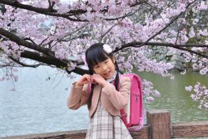 桜の前でハートのポーズをとるピンクのランドセルを背負った女の子