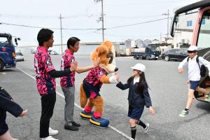 バスから降りてくる子どもたちをタッチで出迎えるセレッソ大阪アンバサダーの男性2人と公式キャラクターのロビーくん