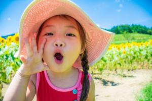 ひまわり畑の前で、口の横に右手を添え「おーい」と呼びかける麦わら帽子を被った女の子